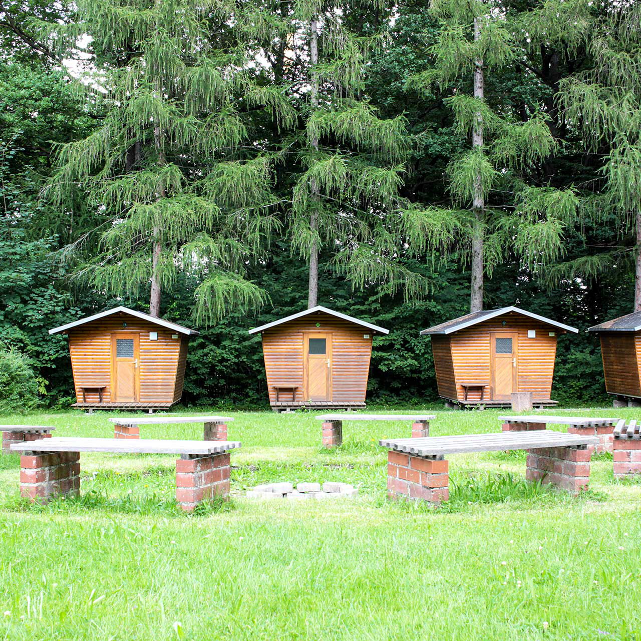 Die Lagerfeuerstelle auf der großen Wiese mit acht festen Sitzbänken und im Vordergrund stehen vier Holzhäuschen.
