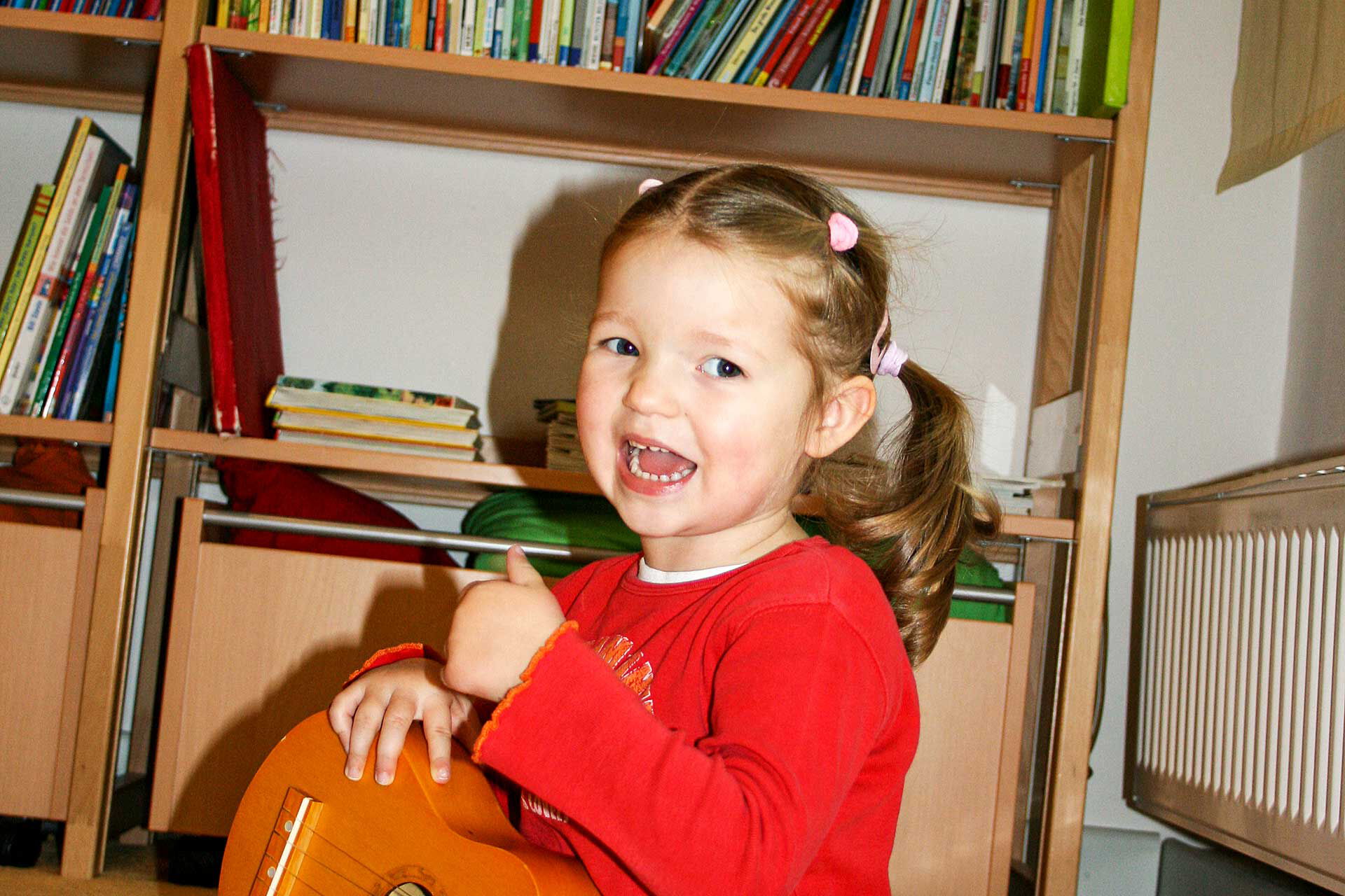Ein Mädchen sitzt auf dem Boden und spielt mit einer kleinen Holzgitarre. Im Hintergrund steht ein Regal mit Büchern.