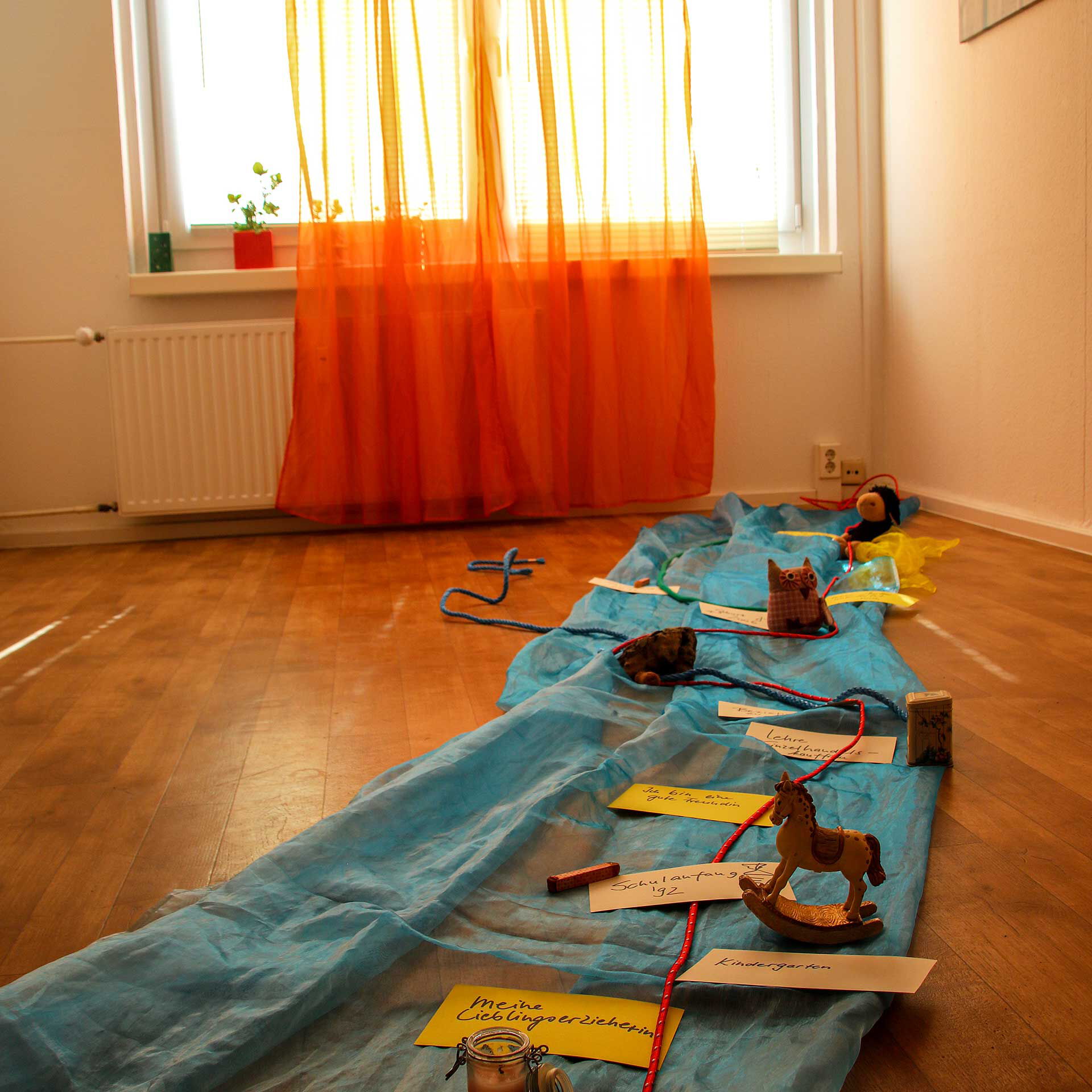 Auf dem Fußboden des Zimmers liegt ein selbstgebastelter Zeitstrahl des Lebens aus Seilen, Stricken, Spielzeugfiguren und blauen Stoff.