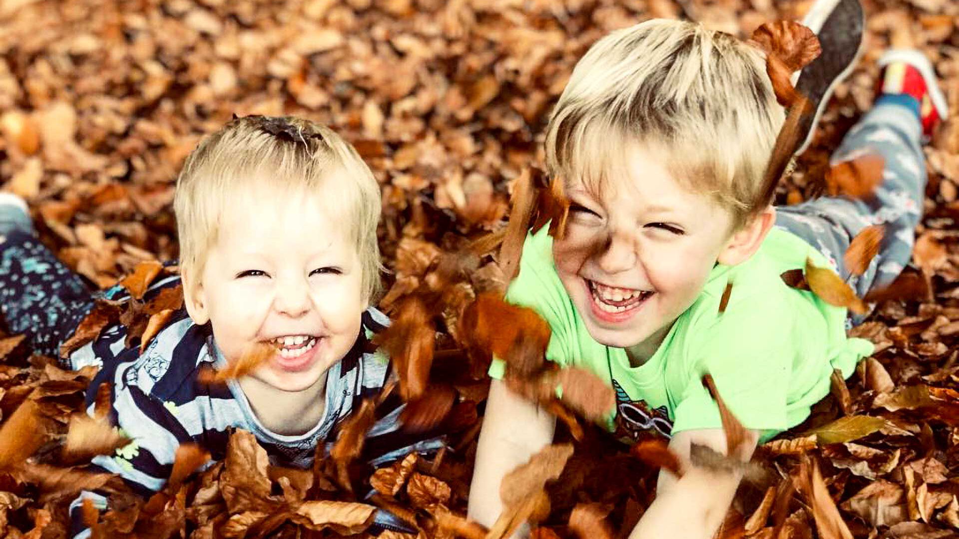 Zwei Kinder liegen lachend auf dem Waldboden im Laub.