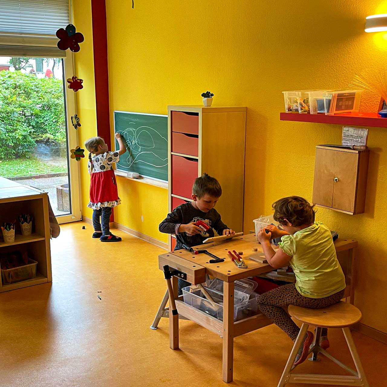 Eine Werkbank steht an der Wand in einem Zimmer und zwei Kinder sitzen auf Stühlen an der Werkbank. Ein Mädchen malt mit Kreide an eine Wandtafel.