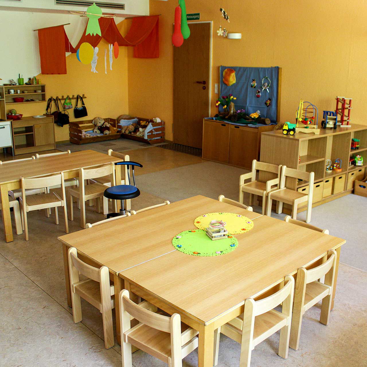 Ein Gruppenzimmer mit zwei großen Tischen und Stühlen. An den Wänden stehen Regale und eine kleine Spielzeugküche.