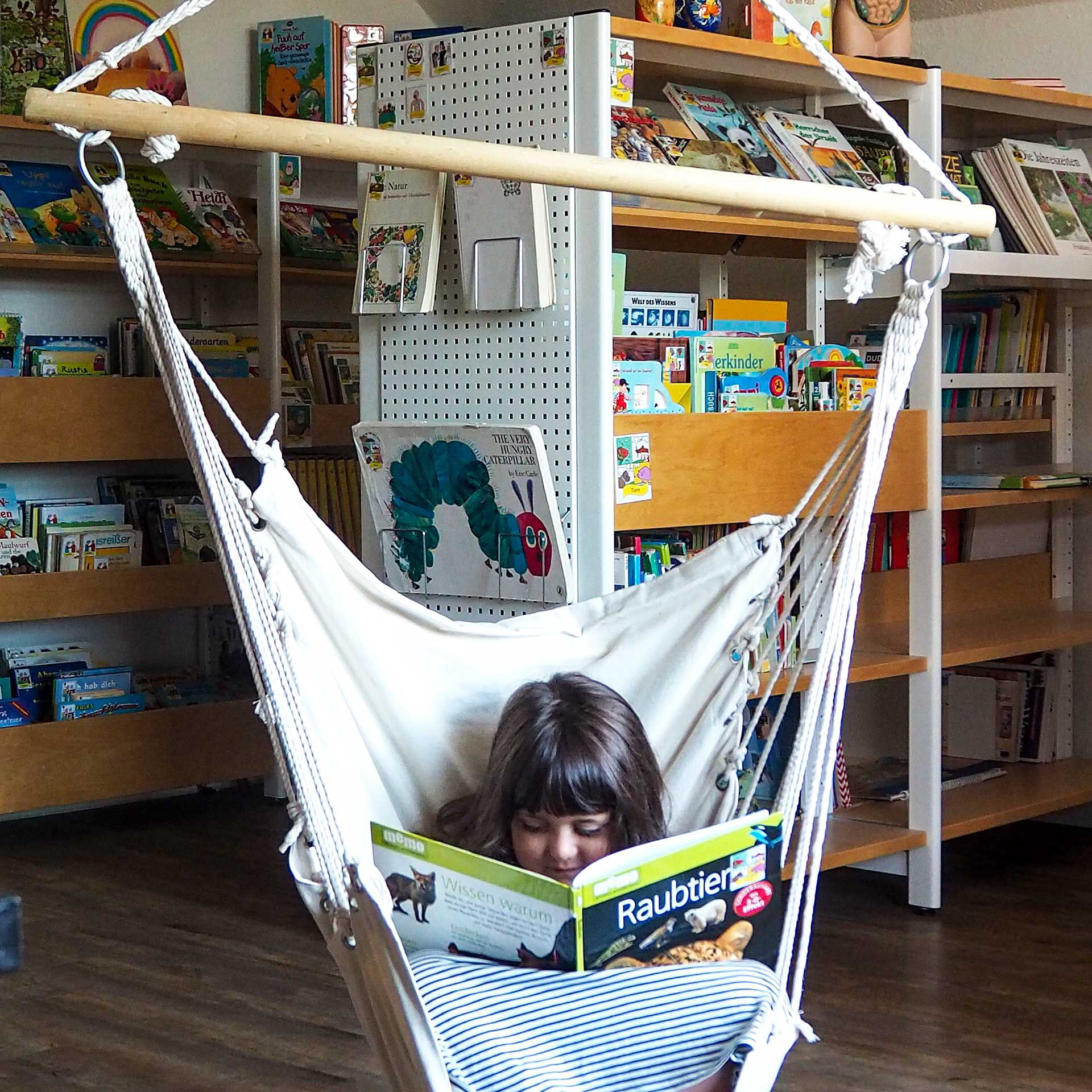 Ein Kind sitzt in einem Hängesessel und schaut sich ein Buch ein. Im Hintergrund stehen Regale mit Büchern.