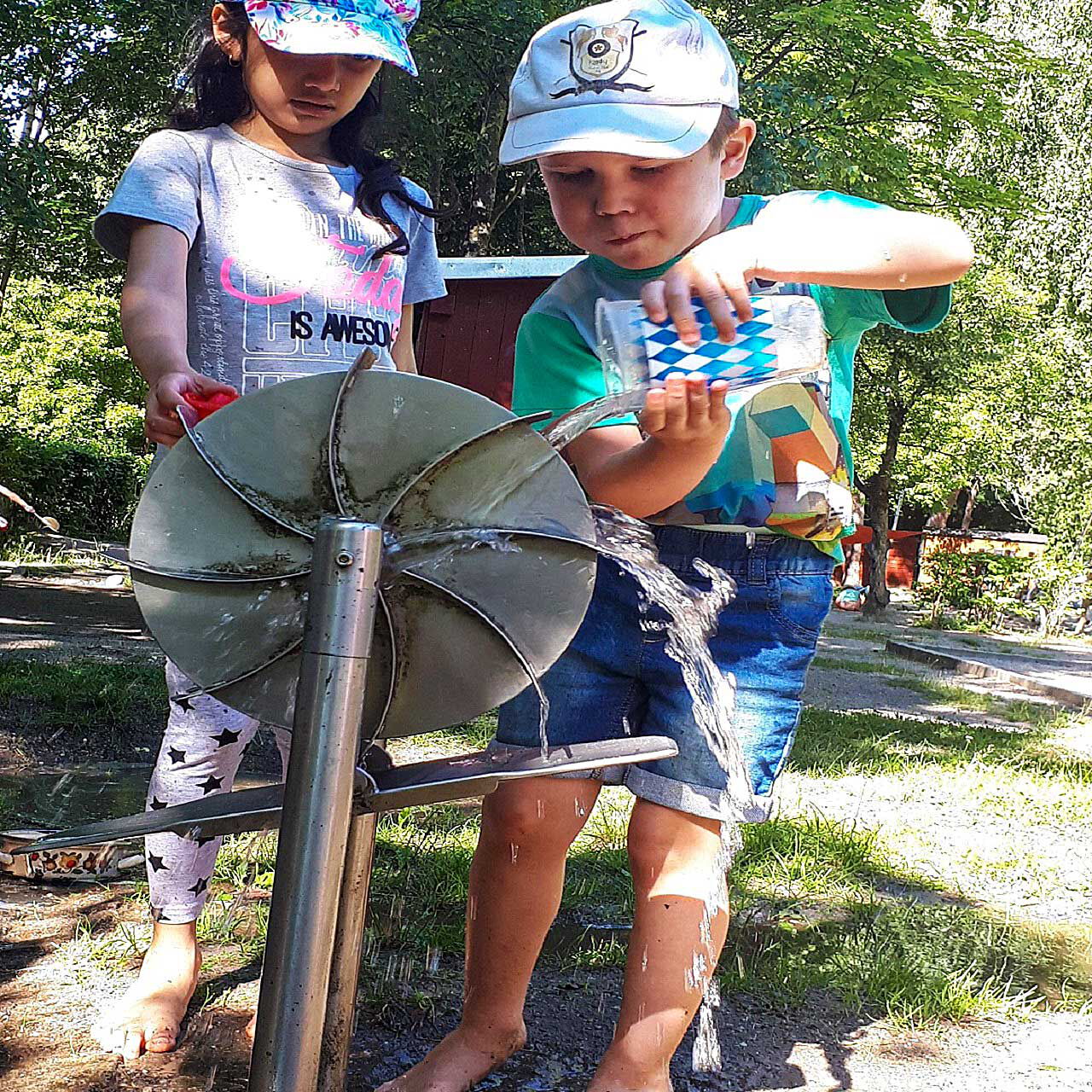 Ein Wasserrad aus Metall im Gartenbereich. Zwei Kinder spielen mit dem Wasserrad und mit Wasser befüllte Becher.