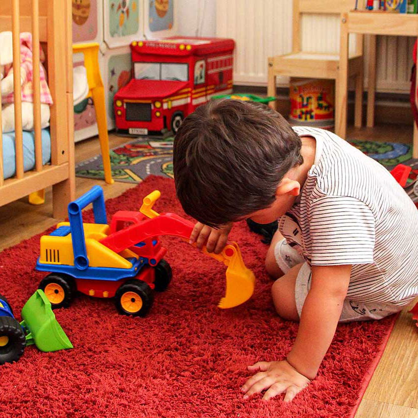 Im Spielbereich sitzt ein Kleinkind auf einem Teppich und spielt mit einem Bagger. Im Zimmer stehen ein Kinderbett und viele Spielsachen.
