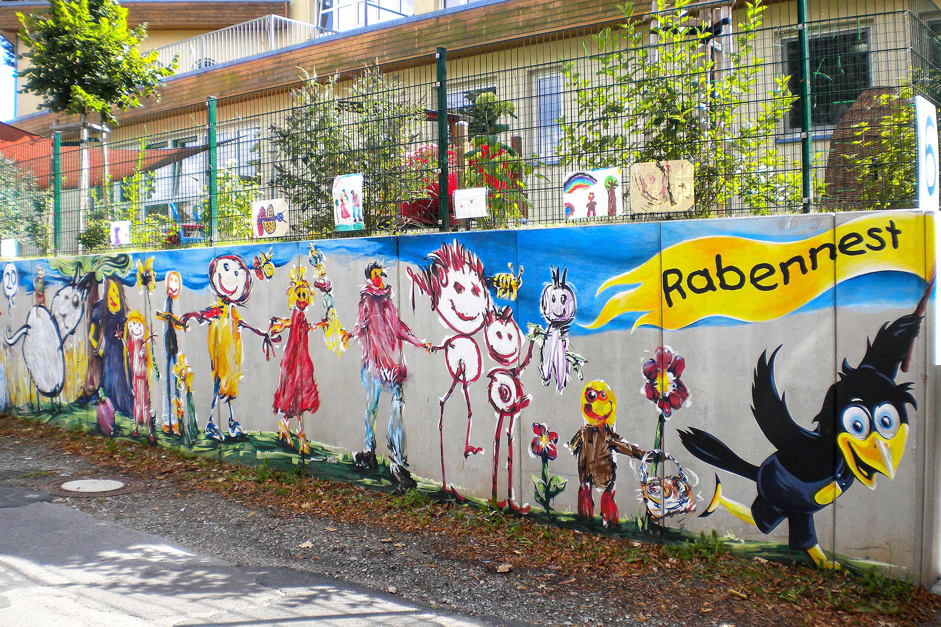 Die bunte Stützmauer zur Kieselhausenstraße ist mit vielen bunten Kinderbildern gestaltet worden. Ein Rabe hält eine gelbe Fahne mit der Aufschrift Rabennest in der Hand.