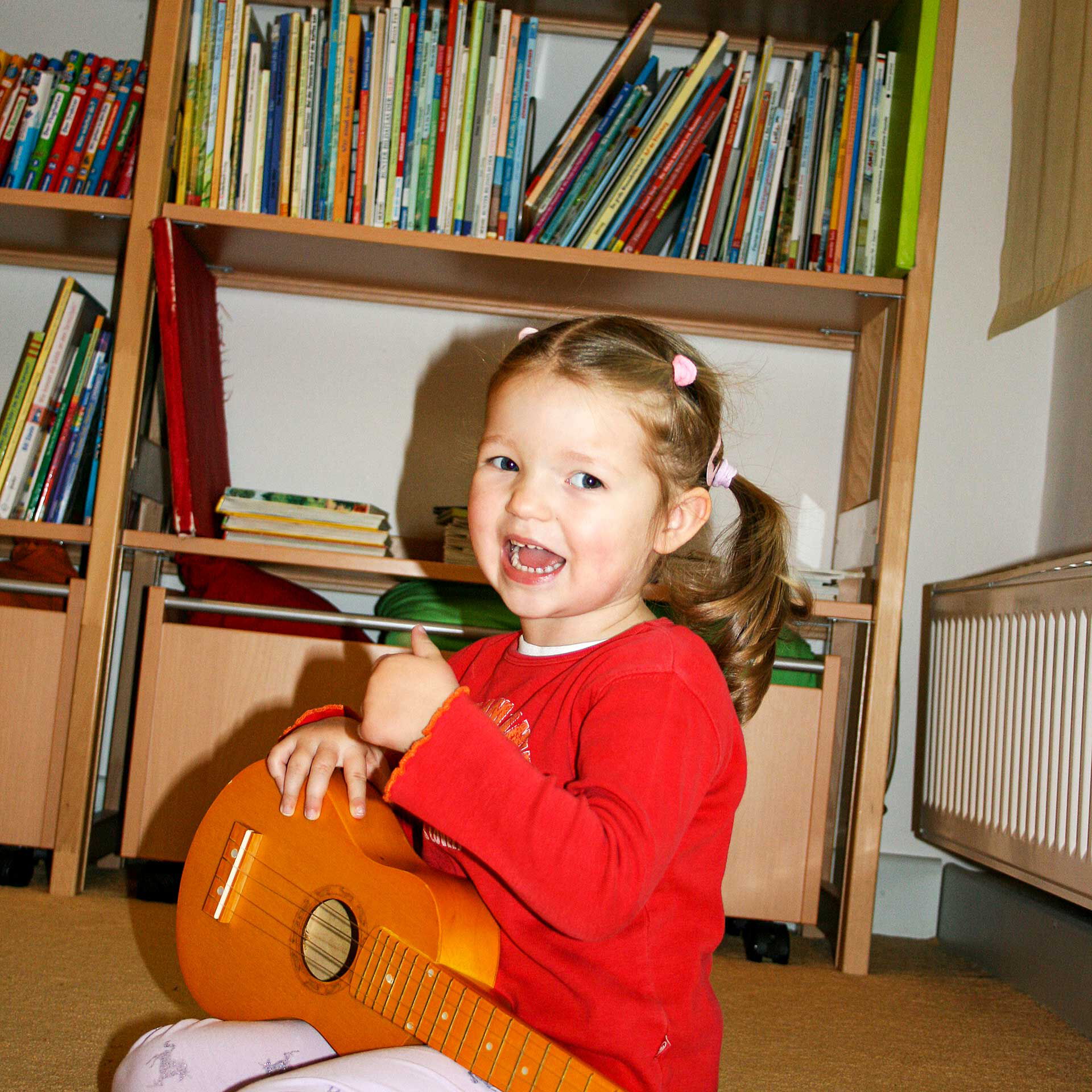 Ein Mädchen sitzt auf dem Boden und spielt mit einer kleinen Holzgitarre. Im Hintergrund steht ein Regal mit Büchern.