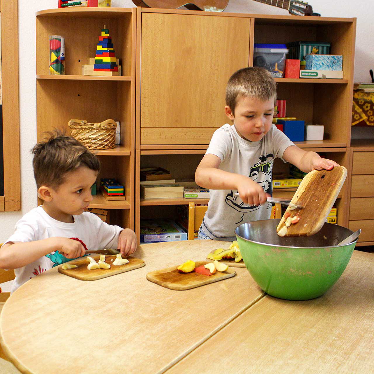 Zwei Jungs sitzen an einem Tisch und bereiten einen Obstsalat zu. Ein Junge schneidet einen Apfel und der andere schüttet seine Apfelstücke in eine große Schüssel.