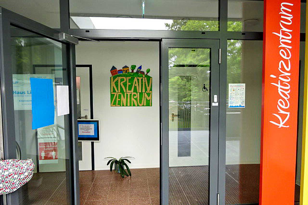 Der Eingangsbereich des Kreativzentrums bestehend aus einer vollverglasten grauen Eingangstür, einem Schriftzug Kreativzentrum auf der rechten Seite und einem selbst gemalten Bild des Kreativzentrums im Bildmittelpunkt.