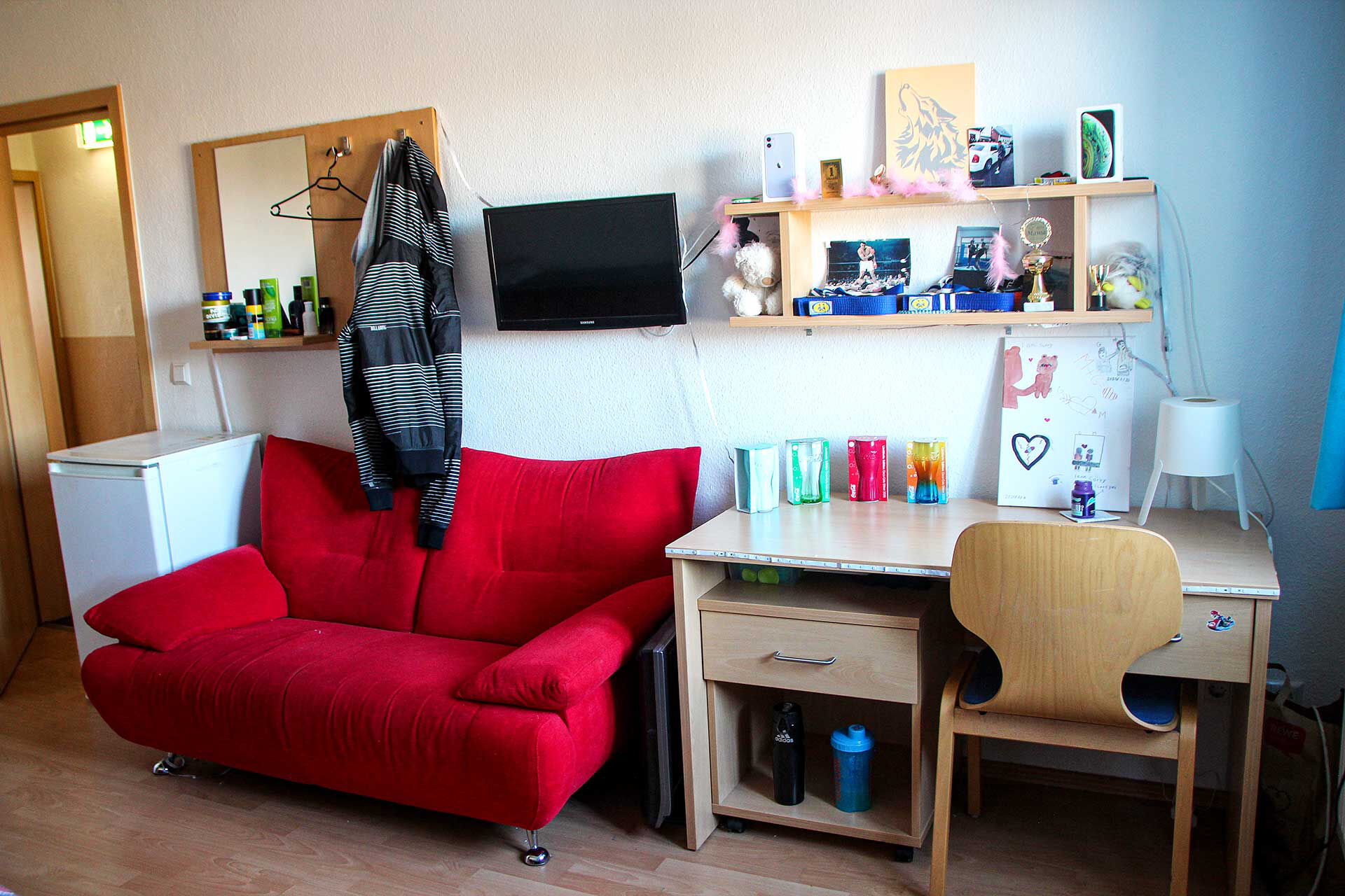 Ein Jugendzimmer mit Schreibtisch, rotem Zweisitzer, Kühlschrank und Fernseher an der Wand.