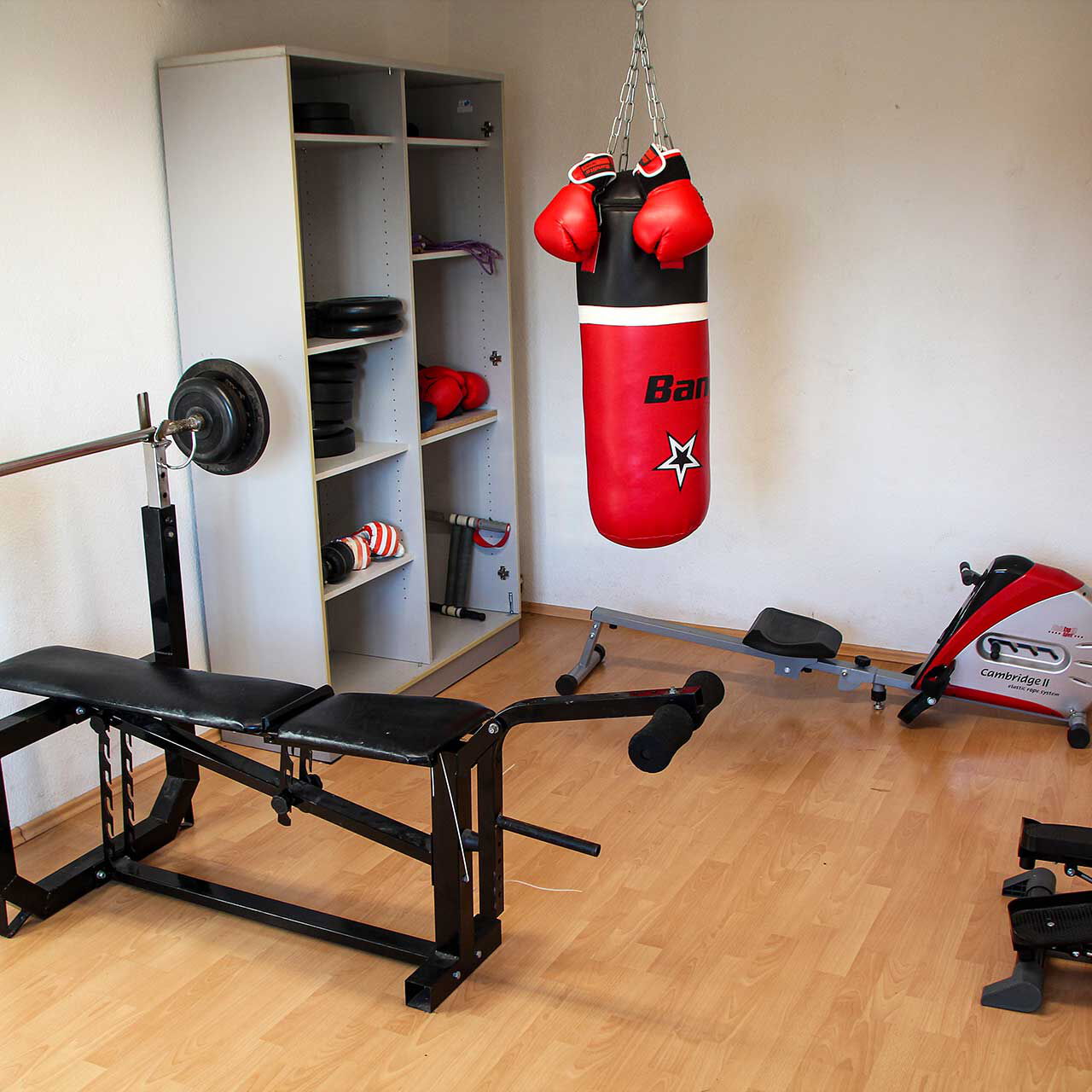 Eine Sport- und Fitnessraum mit einer Hantelbank, einem Boxsack, Schrank mit Gewichten, Stepper und Rudergerät.