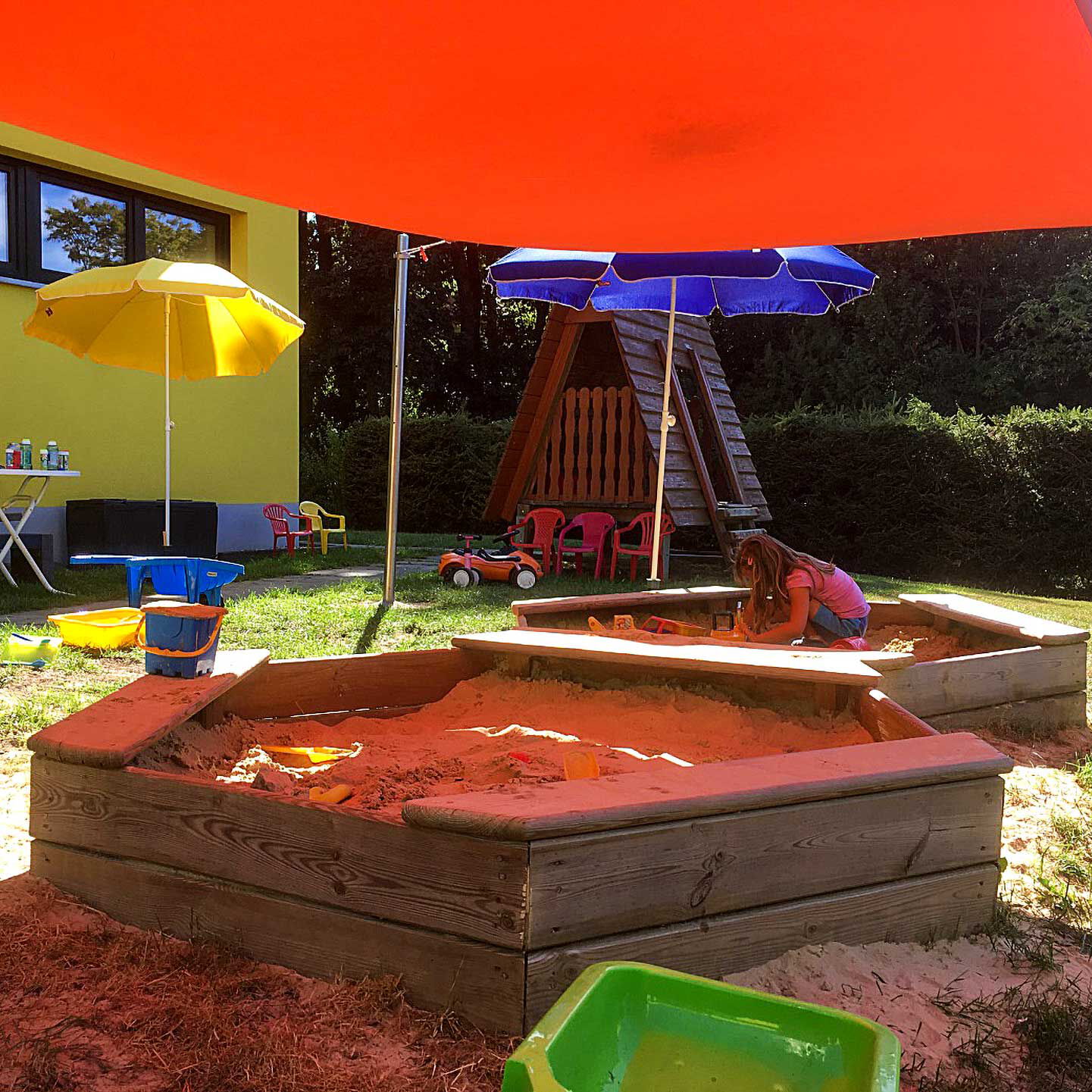 Im Außenbereich sind zwei Sandkästen mit Sonnenschutz, ein Spielhaus und Kinderspielzeuge zu sehen.