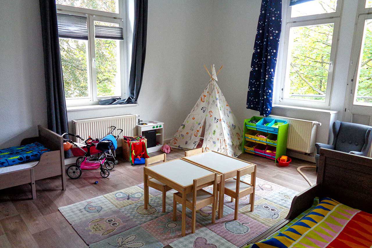 Eine Spielecke in einem Kinderzimmer mit Regal, Tipi, Kinderküche, Puppenwagen und zwei Kinderbetten.