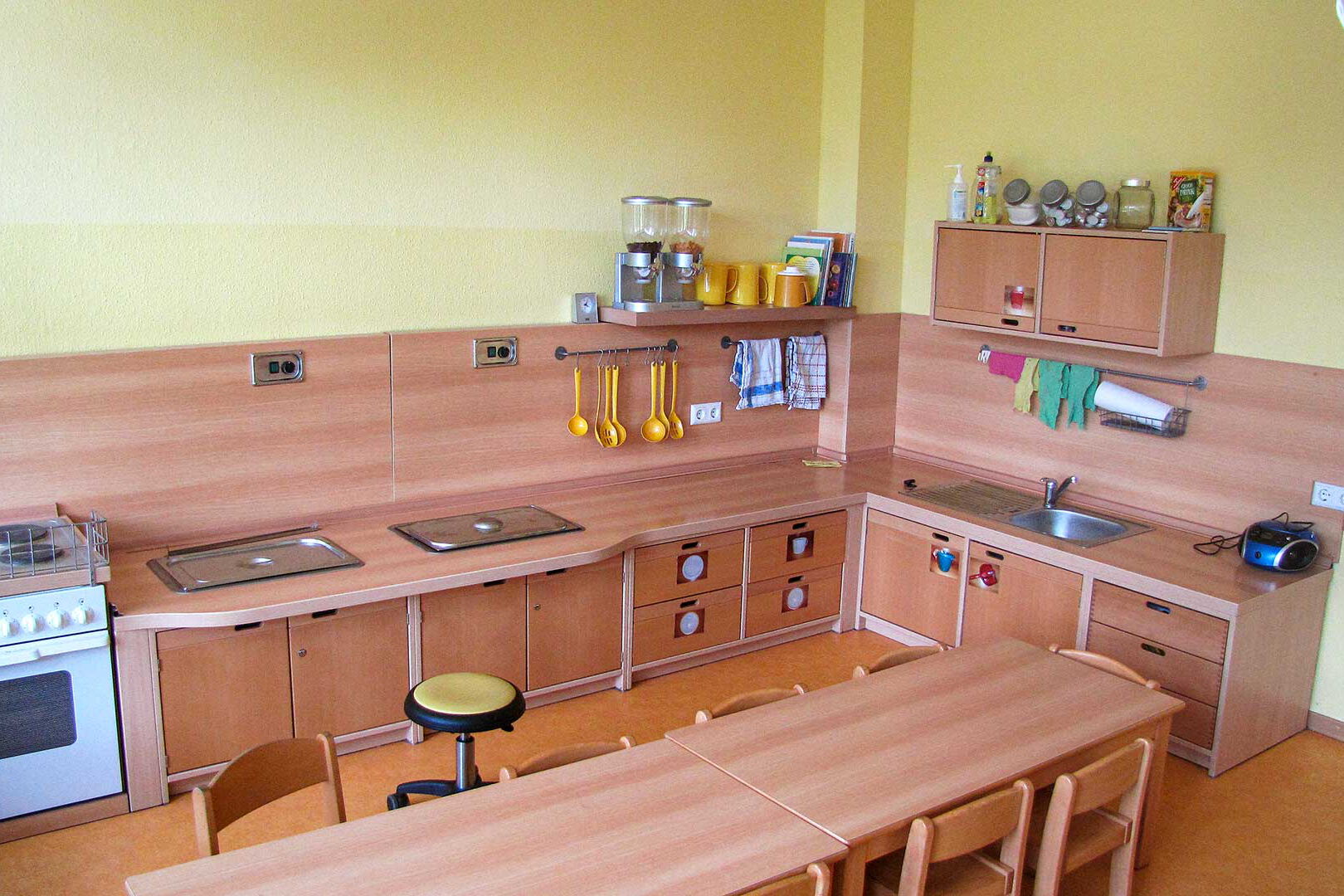 Die Kinderküche mit einer großen Küchenzeile, einem Herd, Waschbecken und zwei langen Tischen mit Stühlen.