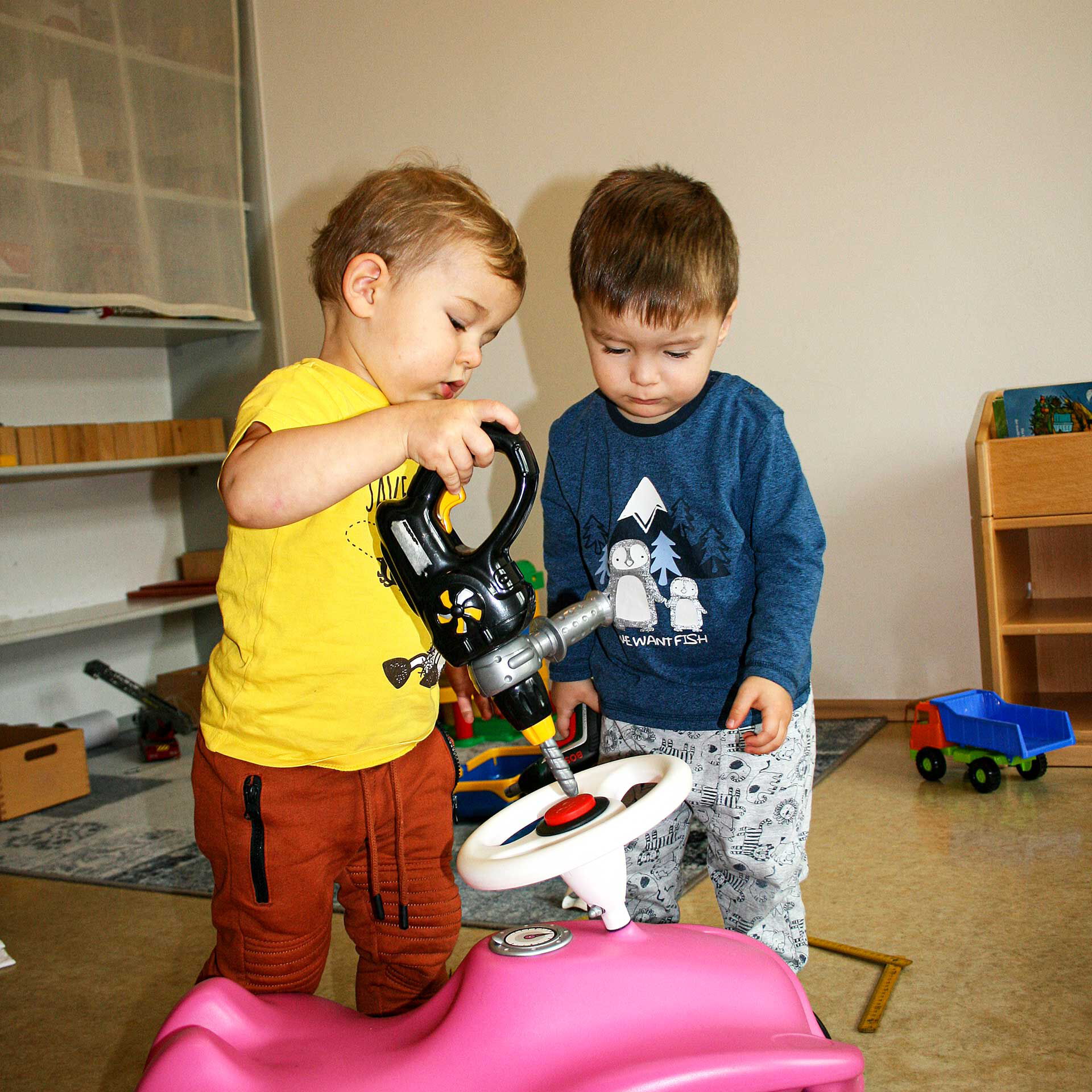 Ein Spielzimmer mit verschiedenen Autos und Bausteinen in dem zwei Kleinkinder zusammen mit einem Bobby-Car spielen.