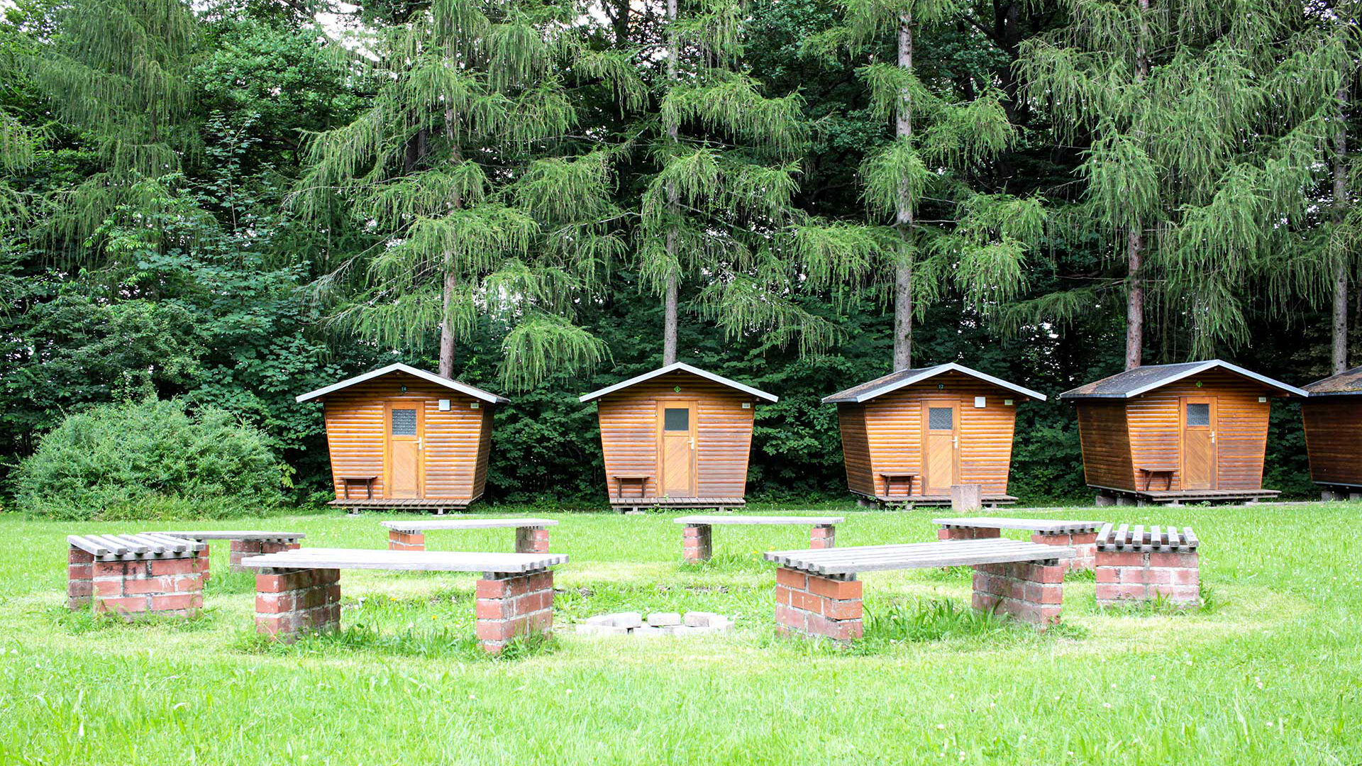 Die Lagerfeuerstelle auf der großen Wiese mit acht festen Sitzbänken und im Vordergrund stehen vier Holzhäuschen.