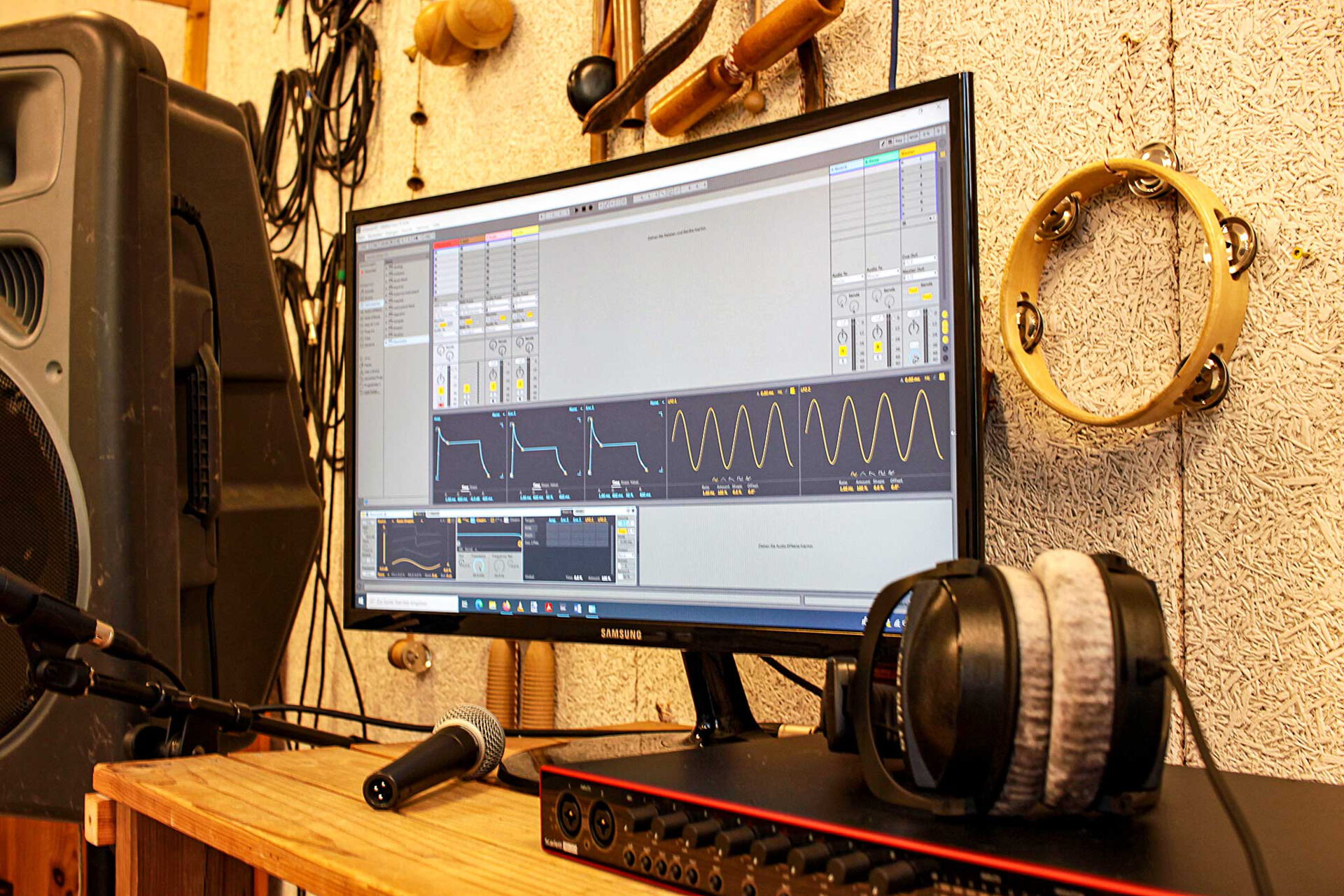 Ein Bildschirm mit Musiksoftware, Kopfhörern und Mischpult. An der Wand hängen mehrere Musikinstrumente.