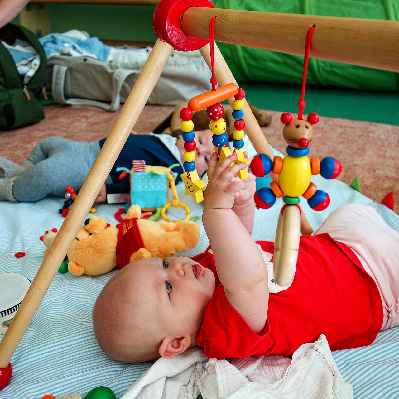 Zwei Babys liegen rücklinks auf einer Decke auf dem Fußboden und greifen nach verschiedenen Spielzeugen.