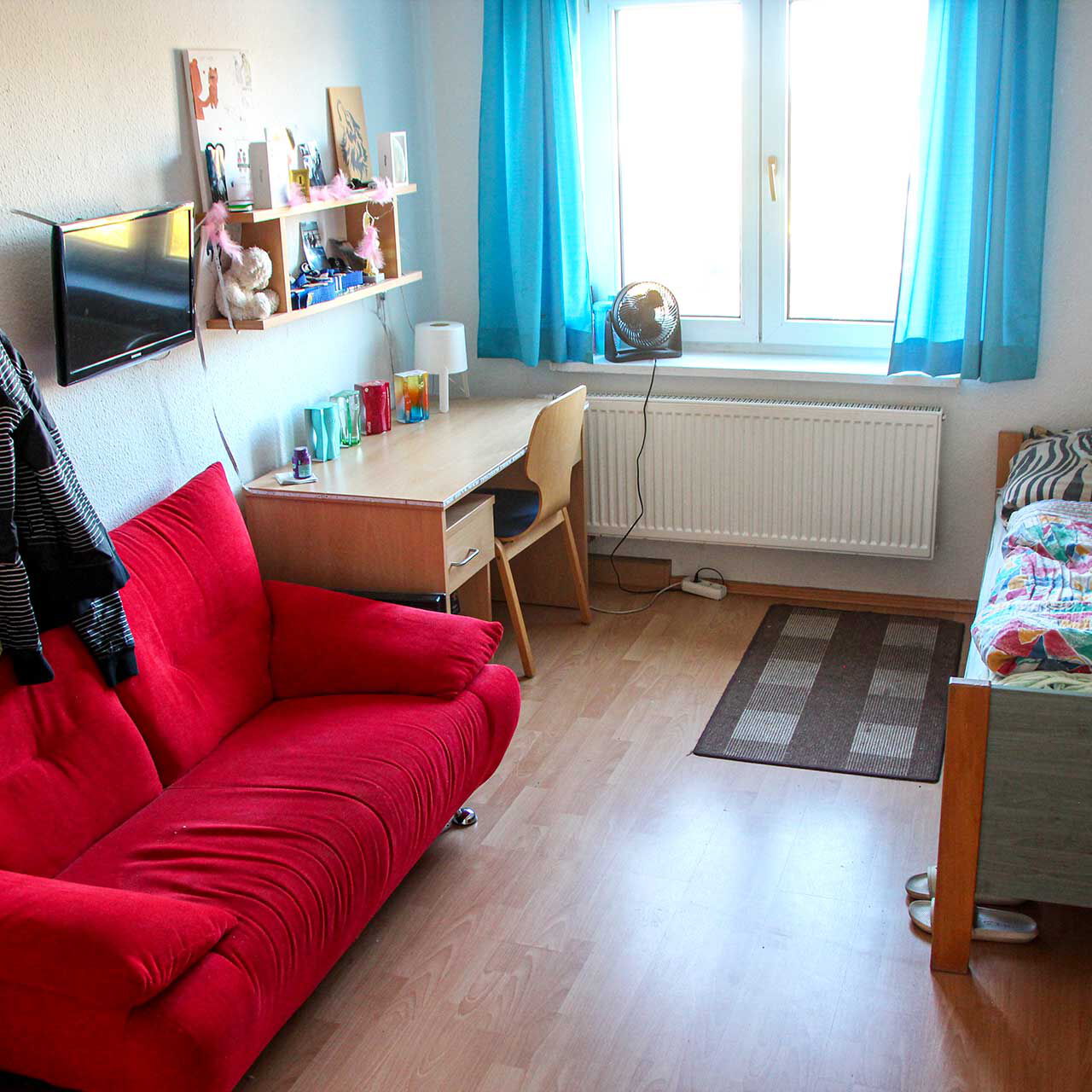 Ein Jugendzimmer mit Bett, Schreibtisch, Kühlschrank, rotem Zweisitzer und einem Fernseher an der Wand.